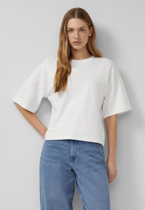 Jonge vrouw met lang blond haar die een los wit shirt met korte mouwen en lichtblauwe spijkerbroek draagt, staand tegen een grijze muur.