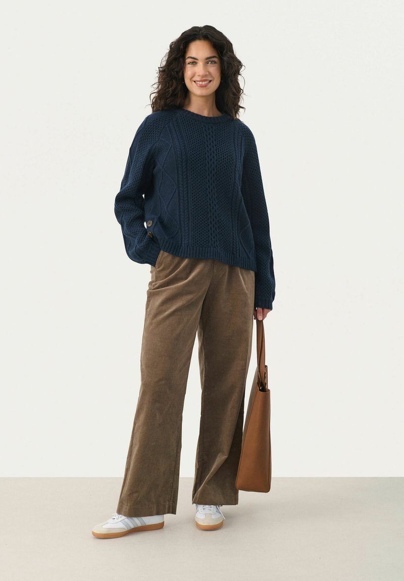 Marineblauer Strickpullover mit strukturiertem Muster, lockere Passform; braune Cordhose, weiße und graue Sneakers und braune Tragetasche.