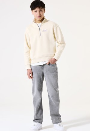 Sudadera cremosa con cremallera y cuello alto, combinada con jeans gris claro y zapatillas blancas, con un ajuste casual y un logo minimalista.
