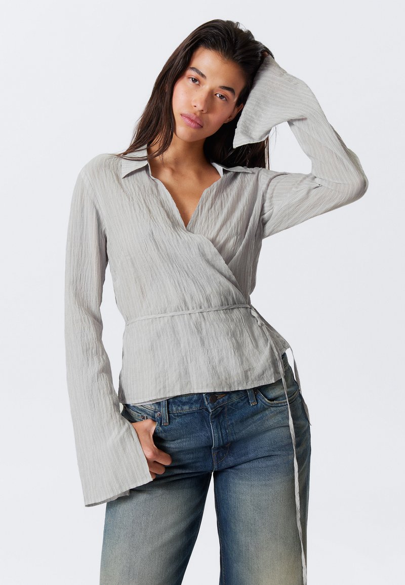 Blouse à enroulement gris clair avec une finition texturée, col pointu et manches longues évasées, associée à un jean bleu délavé.