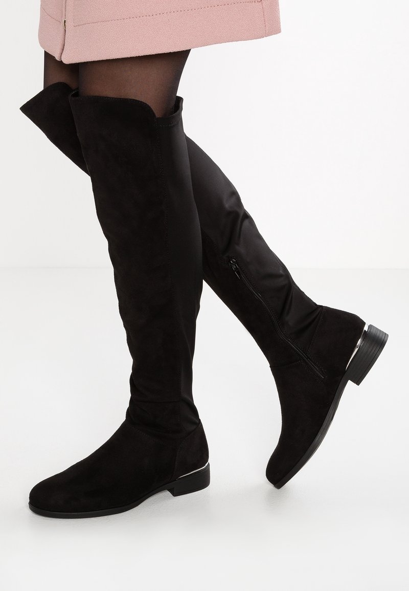 Botas planas negras sobre la rodilla con cremallera lateral, usadas con medias negras transparentes y una falda rosa sobre un fondo blanco.