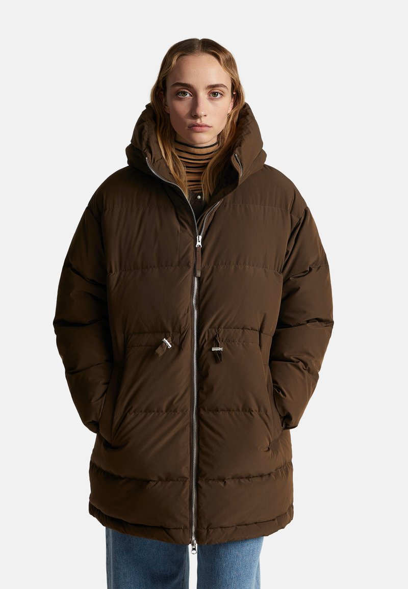 Hnědá péřová bunda s kapucí, s předním zipem, prošívaný design a přední kapsy. Nosená přes pruhované tričko a modré džíny.