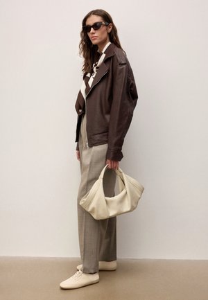 Kobieta ubrana w oversize'ową brązową skórzaną kurtkę, beżowe spodnie w kratę, białe trampki, brązowo-biały szalik, czarne okulary przeciwsłoneczne, trzymająca dużą kremową torbę.