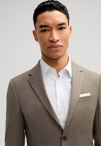 Mężczyzna z krótkimi kręconymi włosami, ubrany w beżową marynarkę na białej koszuli, na jasnym, jednolitym tle.