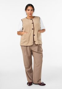 Beige fleece vest med brun kant og knapper, med to forlommer. Kombineret med lysbrune bukser og maroon sko.