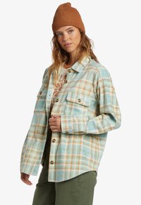 Flannel shirt in blue, green, and orange plaid. Features a button-up front, two chest pockets, and a relaxed fit. Accompanied by a brown beanie.
