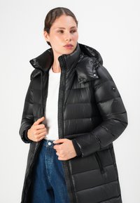 Vrouw in een zwarte, gewatteerde jas met rits en hoge kraag, voorzien van een verstelbare capuchon, gecombineerd met blauwe jeans en een witte top.