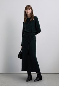 Pletený dvoudílný komplet v tmavě zelené barvě, zahrnující top s dlouhým rukávem a výstřihem a přiléhavou sukni doplněnou ozdobným páskem ze šňůrky.