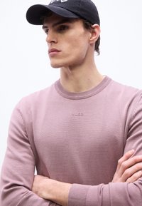 Νέος άνδρας με σταυρωμένα χέρια, φορώντας πουλόβερ μωβ απόχρωσης με το λογότυπο "HUGO" και μαύρο καπέλο μπέιζμπολ, κοιτάζοντας στο πλάι με φόντο ένα απλό υπόβαθρο.