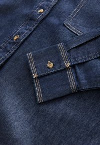 Dunkelblaues Denimhemd mit hellbraunen Nähten, ausgestattet mit einem Button-Down-Kragen und einer einzelnen Tasche am linken Ärmel.