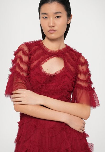 Raudona tiulio suknelė su raukšlėtėmis, trumpomis permatomomis rankovėmis ir rakto skylutės pjūviu iškirpte. Lygus audinys ir lengvas dizainas.