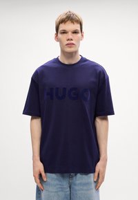 Tamsiai mėlynas medvilninis marškinėliai su dideliu, ryškiu "HUGO" logotipu tamsesnėje atspalvyje. Marškiniai turi laisvą siluetą ir trumpas rankoves.