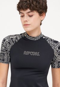 Short-sleeve black rash guard with patterned sleeves featuring seashell designs; "RIPCURL" logo in white center; smooth, stretchy fabric.
