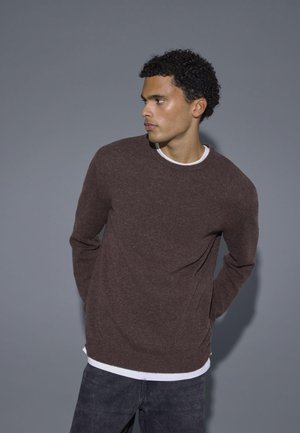 Suéter marrón de punto con mangas largas y cuello redondo, llevado sobre una camisa blanca. Textura rica con un sutil patrón acanalado.