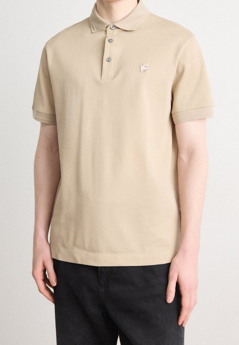 Homme portant un polo à manches courtes beige avec un petit logo brodé sur la poitrine et un pantalon noir, sur un fond uni.