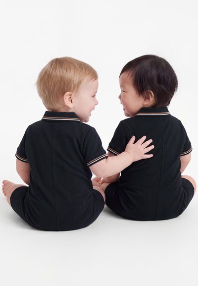 Due bambini seduti vestiti con abiti neri coordinati, uno sorride e tocca delicatamente la schiena dell'altro.