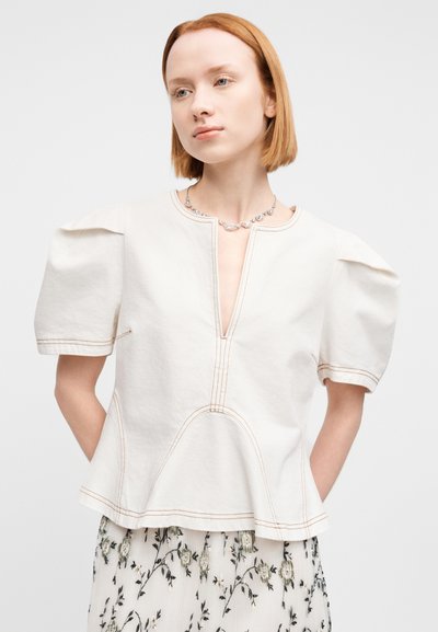 Μπλούζα σε κρεμ απόχρωση από βαμβάκι με φουσκωτά μανίκια, βαθιά V λαιμόκοψη και εμφανείς ραφές. Φορεμένη πάνω από φούστα με λουλουδάτο μοτίβο.