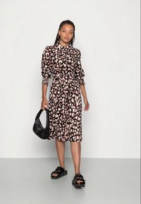 Vestido de padrão de leopardo preto e rosa com mangas longas e volumosas e cintura cinzada, combinado com sandálias pretas e uma mala de ombro preta.