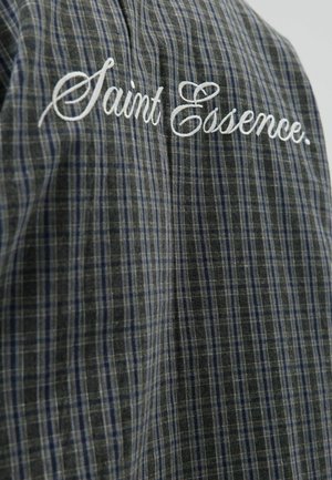 Plaid shirt in dark gray and blue, featuring white embroidered text "Saint Essence." Made of lightweight fabric with a soft texture.