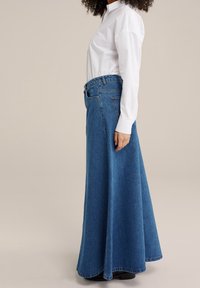 Džínová maxi sukně v modré barvě s vysokým pasem a rozšířenou siluetou. Obsahuje klasický design s pěti kapsami a prošívané detaily na lemu.