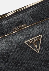 Czarna torebka kopertówka z fakturowanej skóry z złotym zamkiem błyskawicznym i metalowymi elementami, z trójkątnym złotym logo "GUESS". Wewnątrz znajduje się kieszeń zapinana na zamek.