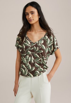 T-shirt à manches courtes avec un col en V, présentant une base marron avec des motifs de feuilles verts et crème. Tissu doux avec une coupe ample, associé à un pantalon crème.