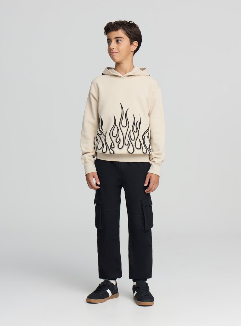 Beżowa bluza z kapturem z czarnym wzorem płomieni na dole, zestawiona z czarnymi spodniami cargo i czarnymi trampkami z białymi akcentami. Gładki materiał, swobodny krój.