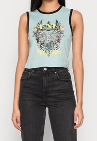 Ärmlös ljusblå crop top med svart kant, utsmyckad med en intrikat blommönster och vingad design samt gul text, matchad med högmidjade svarta jeans.