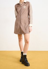 Braunes Reißverschlusskleid mit cremefarbenem Kragen und Ärmeln mit drei Streifen, mit einem Logo. Kombiniert mit schwarzen Schnürstiefeln und weißen Socken.