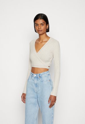 Top envolvente com textura canelada em bege claro, com mangas longas, decote em V e um design cropped, combinado com jeans de ganga azul claro de cintura alta.