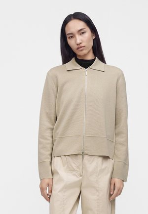 Femme aux longs cheveux noirs portant un pull beige zippé par-dessus un haut noir et un pantalon beige, devant un fond gris clair uni.
