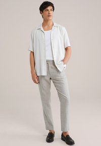 Kurzärmliges weißes Hemd mit Knöpfen, darunter ein weißes Tanktop, graue Nadelstreifenhose, schwarze Slipper und eine Uhr am linken Handgelenk.