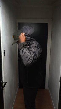 Black puffer jacket with a hood and fleece lining, zippered pockets, worn with a dark cap, in an indoor corridor with neutral walls.
