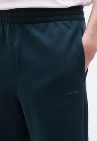 Άνδρας που φοράει σκούρες μπλε φόρμες Calvin Klein με ελαστική μέση και το χέρι του στην δεξιά τσέπη.
