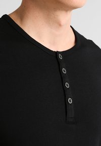 Camisa henley de algodón negro con cuello redondo y tapeta de cinco botones, que presenta una textura suave y un diseño ajustado.