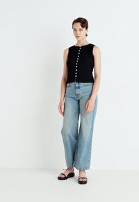 Juodas suknelinis tanktopas su priekinių mygtukų užsegimu, derinamas su šviesiai mėlynomis plačiomis kelnėmis ir juodais plokščiais sandalais. Balta fono spalva, minimalūs aksesuarai.