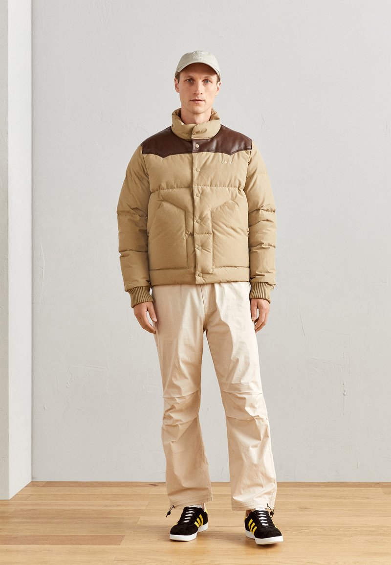 Giacca puffer beige con yoke in pelle marrone sulle spalle, abbinata a pantaloni crema e sneaker nere con strisce gialle.