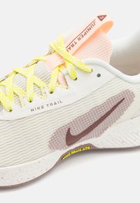 Nike trail løbesko med en tekstureret lys grå overdel, fleksible gule snørebånd, lyserøde accenter og en brun Nike Swoosh-logo.
