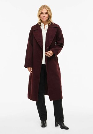 Burgundy long coat made of soft fabric, featuring a wide collar, button closure, and loose fit. Styled over a light shirt and black trousers.