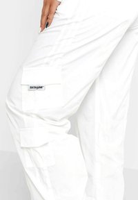 White cargo pants with multiple large pockets and a stitch design. Features a tag with the brand name "SIXTHJUNE." Smooth texture, relaxed fit.