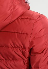 červená prošívaná bunda s horizontálním prošíváním a připevněnou kapucí s bílým a červeným stažením.