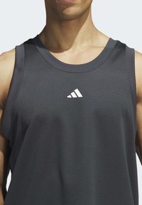 Tmavě šedý sportovní tílko vyrobené z prodyšného síťového materiálu. Má kulatý výstřih, střihové rukávy a bílé logo Adidas na hrudi.