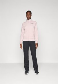 Sweatshirt zippé rose clair avec col montant et logo noir, associé à un pantalon de survêtement gris foncé et des chaussures de sport noires. Texture lisse.