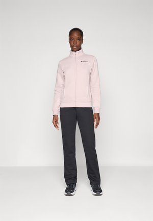 Sweatshirt zippé rose clair avec col montant et logo noir, associé à un pantalon de survêtement gris foncé et des chaussures de sport noires. Texture lisse.