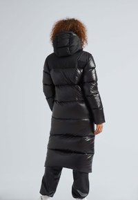Abrigo acolchado negro con capucha, con un exterior brillante y acolchado y rayas horizontales. Marca sutil en la parte posterior. Diseño de largo extendido.