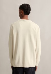 Pull en tricot couleur crème avec une texture côtelée, des manches longues et un col rond. Il présente une coupe décontractée et un design simple.