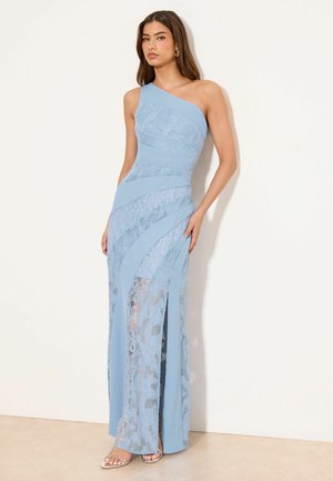 Kobieta w jasno niebieskiej koronkowej sukni na jedno ramię z asymetrycznymi panelami i wysokim rozcięciem z przodu, stojąca przy kremowej ścianie.