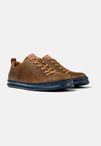 Sneaker in pelle marrone con finitura liscia, punta rotonda, lacci beige e suola in gomma blu scuro. Presenta un tirante sul retro per un indossamento facile.