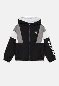 Veste coupe-vent à capuche noire et grise avec une bande blanche et le logo "GUESS" sur la manche, présentée sur fond blanc.