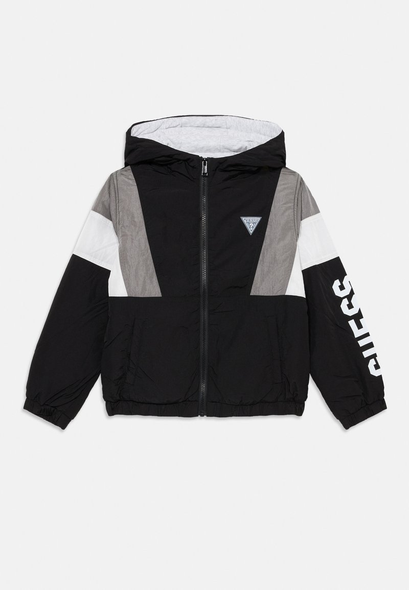 Veste coupe-vent à capuche noire et grise avec une bande blanche et le logo "GUESS" sur la manche, présentée sur fond blanc.